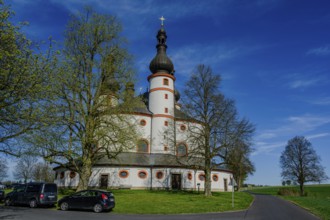 A baroque church with round architecture under a clear blue sky, surrounded by trees and a road,