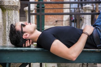 Attractive young man laying down on wood bench in European city, wearing sunglasses