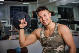 Attractive young athletic man exercising in gym, working out using kettlebell, looking at camera