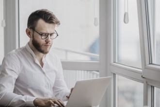 Man in a white shirt and glasses focused on his laptop in a bright room