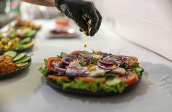 Chef wearing black gloves adding corn grains to a greek salad with tomatoes, red onions, olives,