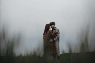 Couple holds each other in a moody open landscape, dressed in trench coats