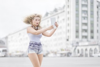 Happy young woman with vintage music headphones around neck taking selfie with tablet PC and