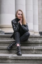 Portrait of young blonde woman with red lips wearing leather jacket on corporate building stairs.