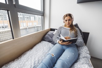 Blonde young caucasian woman lying on her dorm room bed next to a window putting on headphones,