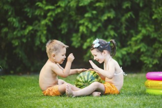 Two children, Caucasian brother and sister, sitting on green grass in backyard of house and hugging