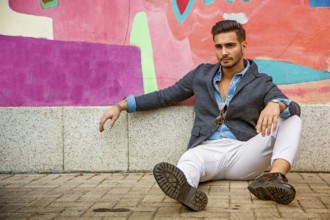 Attractive young man sitting against colorful graffiti wall, looking at camera