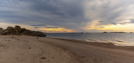 A panorama view of a golden sand beach at sunset with calm ocean water and red granite rocks