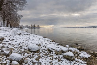 Snow-covered shore of a lake with stones and trees in the background. A peaceful winter landscape