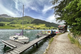 Sailing boat at a jetty on a lake, surrounded by mountains and green nature, under a cloudy sky,