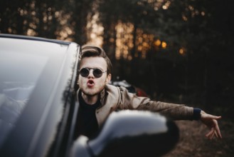 A man in sunglasses leans out of a car, with a forest backdrop reflecting excitement