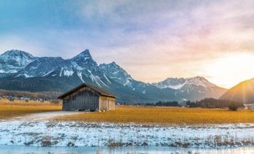 Winter alpine landscape with an aged wooden barn, the Alps mountains, a frozen river at sundown.