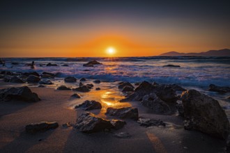 Sunset with rocks in the foreground on the beach. The sea is moving and the sky glows in shades of