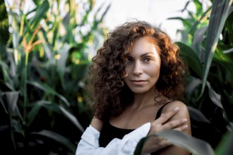 A confident woman with curly hair stands among corn stalks, crossing her arms