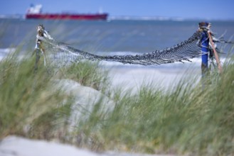 Old, weathered volleyball net in front of grasses on the beach, a ship can be seen in the distance,