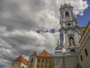 A baroque church tower rising into a cloudy sky, flanked by red-roofed buildings, Dürnstein,