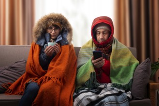 Middle age family in parkas drinking coffee on the sofa while using smartphone in their cold living