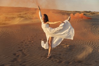 A girl in a fly white dress dances and poses in the sand desert at sunset