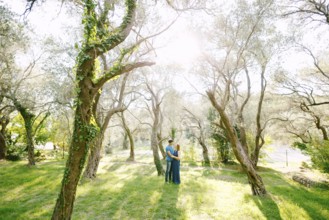 Man hugs pregnant woman in a beautiful park of green olive trees entwined with ivy on a sunny day.