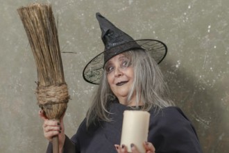 Old witch with white hair and a black hat holding a broom in her hand and a candle in the other