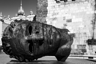 A large metal head sculpture lying on its side near a historical building in black and white,