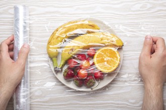 Male hands wrapping assorted different fruit and berries on white plate in plastic clear film top