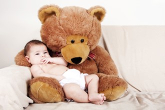 Cute Caucasian Hispanic unisex baby in the arms of a big brown stuffed teddy bear on the couch, New