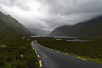 Narrow black-top highway with yellow road markings leads through an overcast mountain valley with