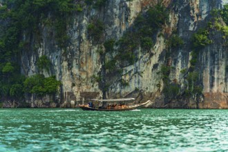 Longboat navigating along a towering cliff with greenery above turquoise water, Cheow Lan Lake,