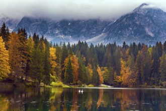 The Dolomites are covered with clouds. Orange and yellow trees are reflected in the green smooth