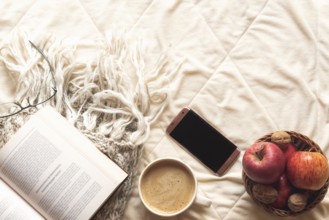 Coziness concept with an open book on a wool shawl, a basket with apples and nuts, a phone and a