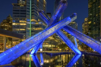 Modern Olympic fire installation at night with blue lighting and surrounding skyscrapers in the