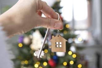 Key and tiny house keychain in hand on background fairy lights Christmas tree. gift for New Year,