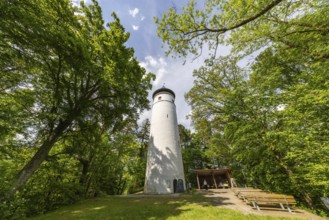 A white observation tower in a park, surrounded by green trees and a small seating area in summer,