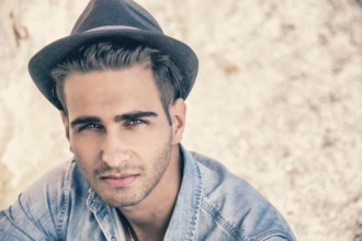 Handsome young, man in denim shirt and fedora hat sitting in pose on sandy beach and looking at