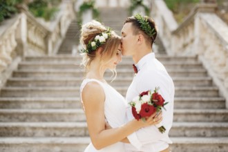 The bride and groom in wreaths stand hugging and kissing on the stairs of the ancient church in