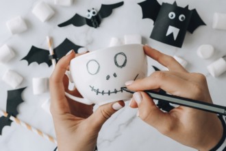 Female hands paint on white cup scary jacks face DIY for kids Halloween home activities Holiday art