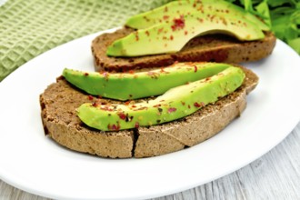 Two slices of rye bread with slices of avocado and pepper on a plate, green cloth, parsley on the