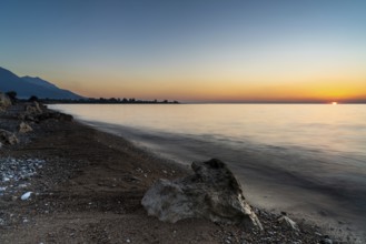 A peaceful sunset over the ocean with silhouette of mountains and pebble beach iwth rock boulders