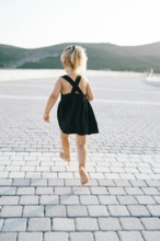 Little girl runs barefoot on a tiled road while hovering in the air. Back view.