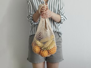 Mesh bag with fruits in female hands. Stylish young woman hold mesh shopping bag on light gray wall