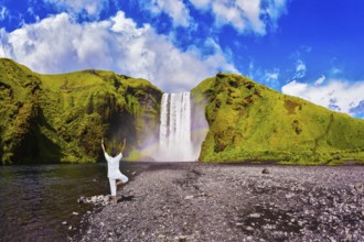 Elderly woman performs asana tree of hatha yoga before falling water. Grand waterfall with rainbow