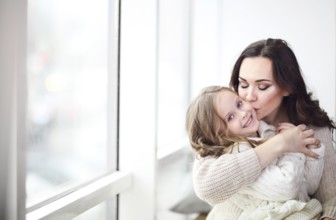 Happy loving family together. Mother and child daughter hugging by the window wearing cozy sweaters