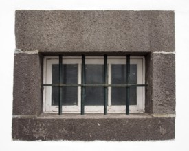A small white wooden window with glass panes covered by steel bars in a large grey stone frame