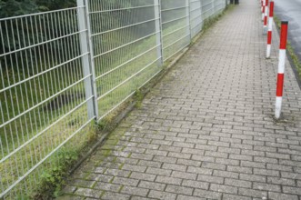 Sidewalk, metal fence on the left, red and white poles on the right