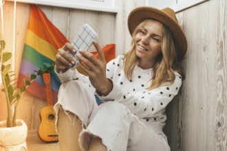 Young millennial hippie woman sitting on balcony using mobile phone. LGBTQ rainbow flag on
