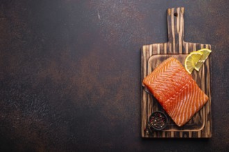 Fresh raw salmon fillet on wooden kitchen cutting board with seasonings and lemon top view on dark