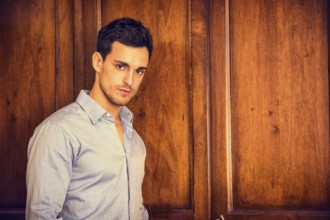 Sexy handsome young man standing wearing shirt in his bedroom against wooden wardrobe door