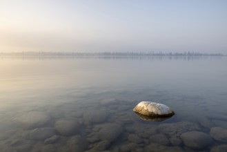 A large stone rests in the calm waters of a misty lake at dawn, harbour, Allensbach,