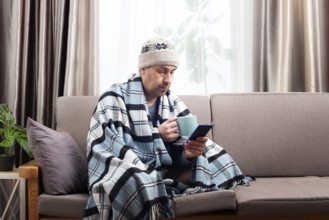 Middle age male model in hat and plaid hold coffee cup sitting on the sofa while using his